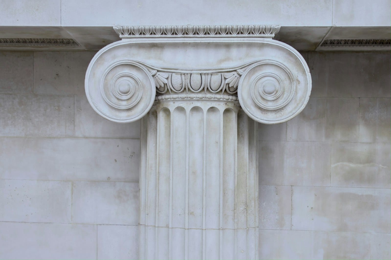 A close-up of an Ionic column capital in weathered stone.