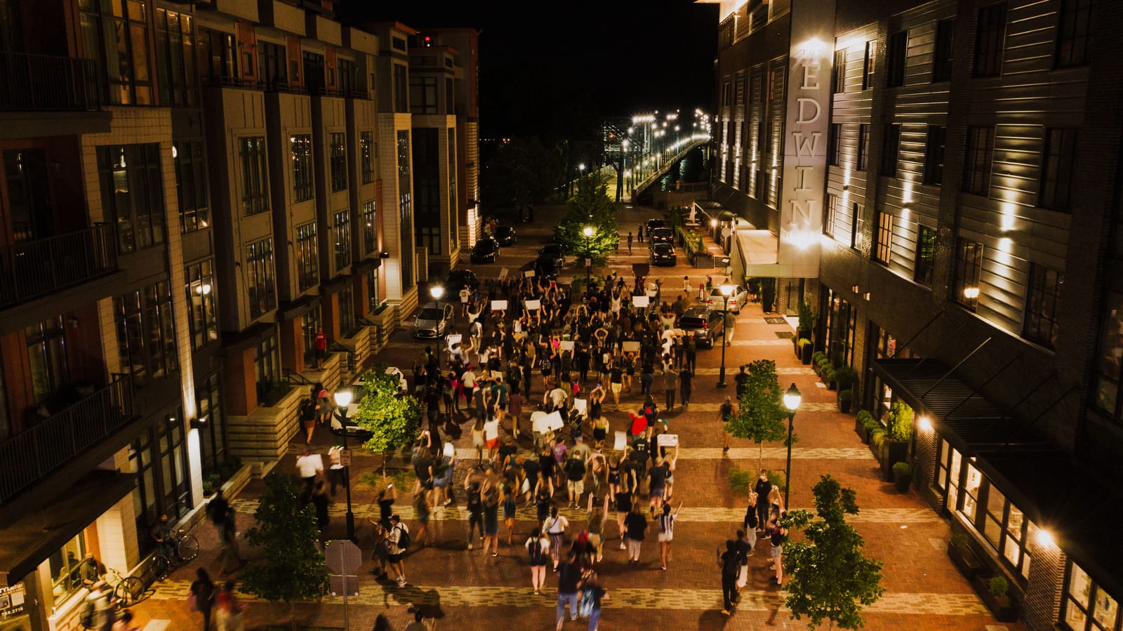 A crowd gathering with placards on a night street, seen from above.