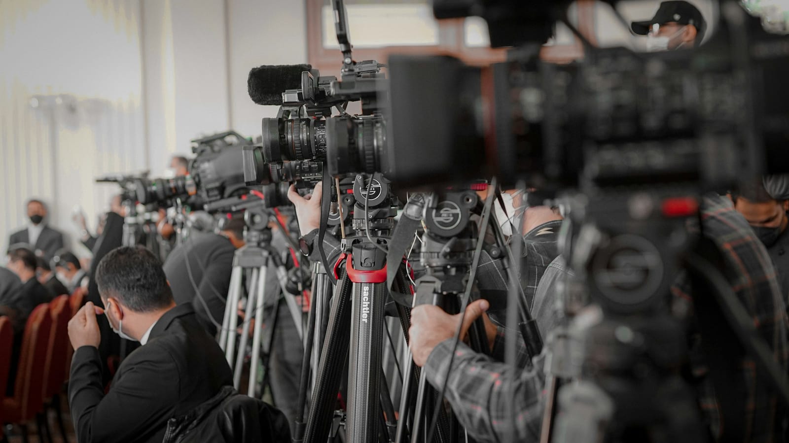 A row of television cameras on tripods trained on a press conference, representing the constant scrutiny of public life.